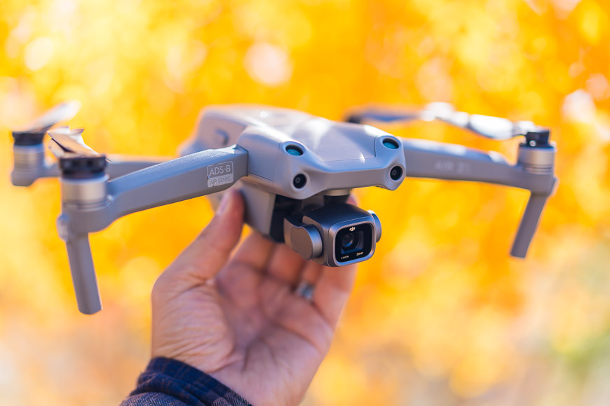 A person holding a small gray drone in front of an autumn tree.