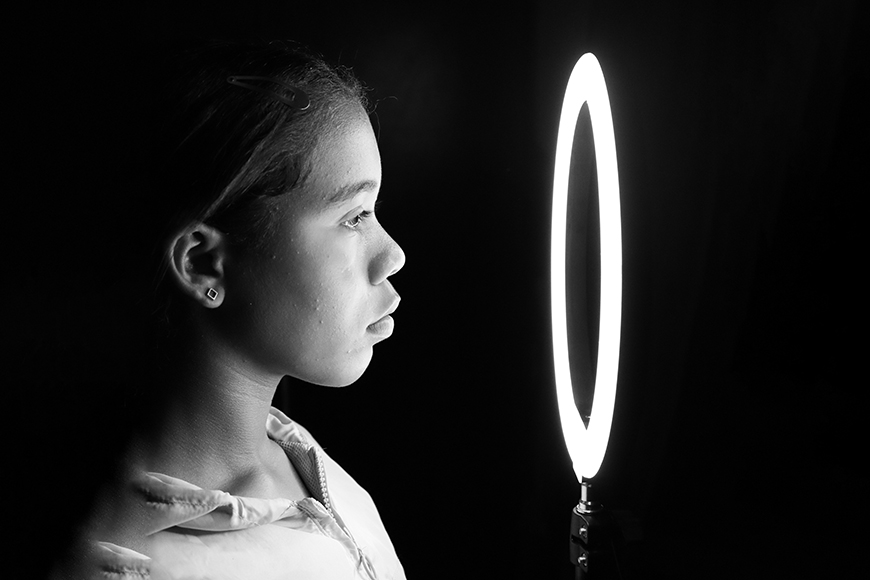 A black and white photo of a girl looking at a ring light.