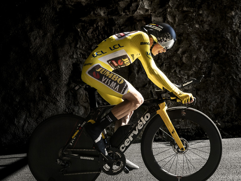 A cyclist in a yellow jersey is riding in front of a cave.