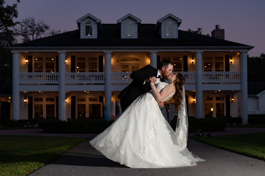 A bride and groom kiss in front of a mansion at dusk.